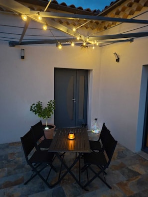 Un patio exterior con una mesa, sillas, luces colgantes, una planta en maceta y una puerta.