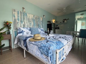 Een slaapkamer met een bed, een bureau, een stoel, een hoed, een fles champagne en een vaas met bloemen.