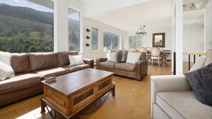 A spacious living room with a large window, a wooden coffee table, and a dining area with a chandelier. - Luxury East Vail Estate w/ Mtn Views & Hot Tub (Vail)