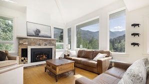 A living room with a fireplace, large windows, a sofa, and a coffee table. - Luxury East Vail Estate w/ Mtn Views & Hot Tub (Vail)