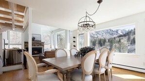 A dining room with a wooden table and chairs, a chandelier, a fireplace, and a large window with a mountain view. - Luxury East Vail Estate w/ Mtn Views & Hot Tub (Vail)