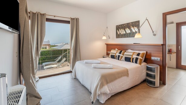 A hotel room with a bed, a view of the outdoors, a painting on the wall, and a lamp. - Modern Villa for 9 Guests on the Cliffs of Cala Morell, Menorca (Ciutadella)