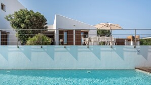 A swimming pool with a clear blue water surface, surrounded by a white wall with a metal railing. There are outdoor seating arrangements with tables and chairs, and a large umbrella providing shade. - Modern Villa for 9 Guests on the Cliffs of Cala Morell, Menorca (Ciutadella)