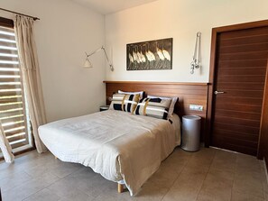 Un dormitorio ordenado con una cama, mesitas de noche, una lámpara de pared, una puerta y un cubo de basura.