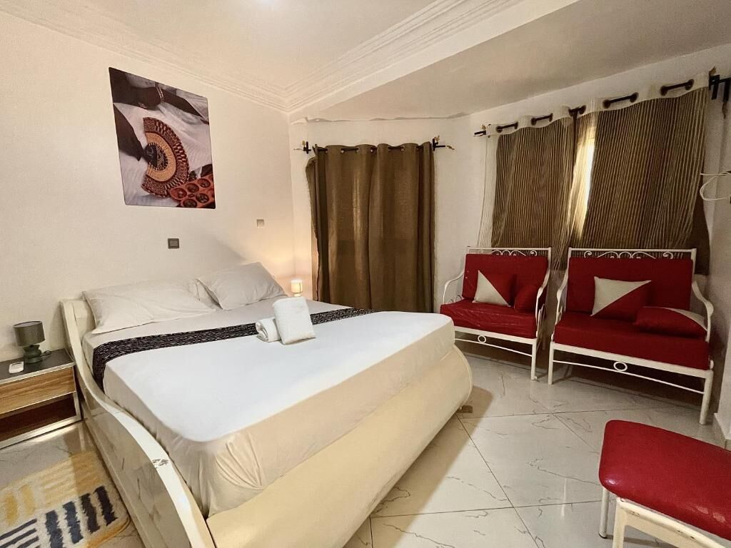 Un dormitorio con una cama, un sofá rojo, un banco rojo, cortinas y un cuadro colgado en la pared.