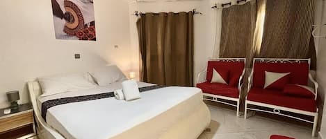 Un dormitorio con una cama, un sofá rojo, un banco rojo, cortinas y un cuadro colgado en la pared.