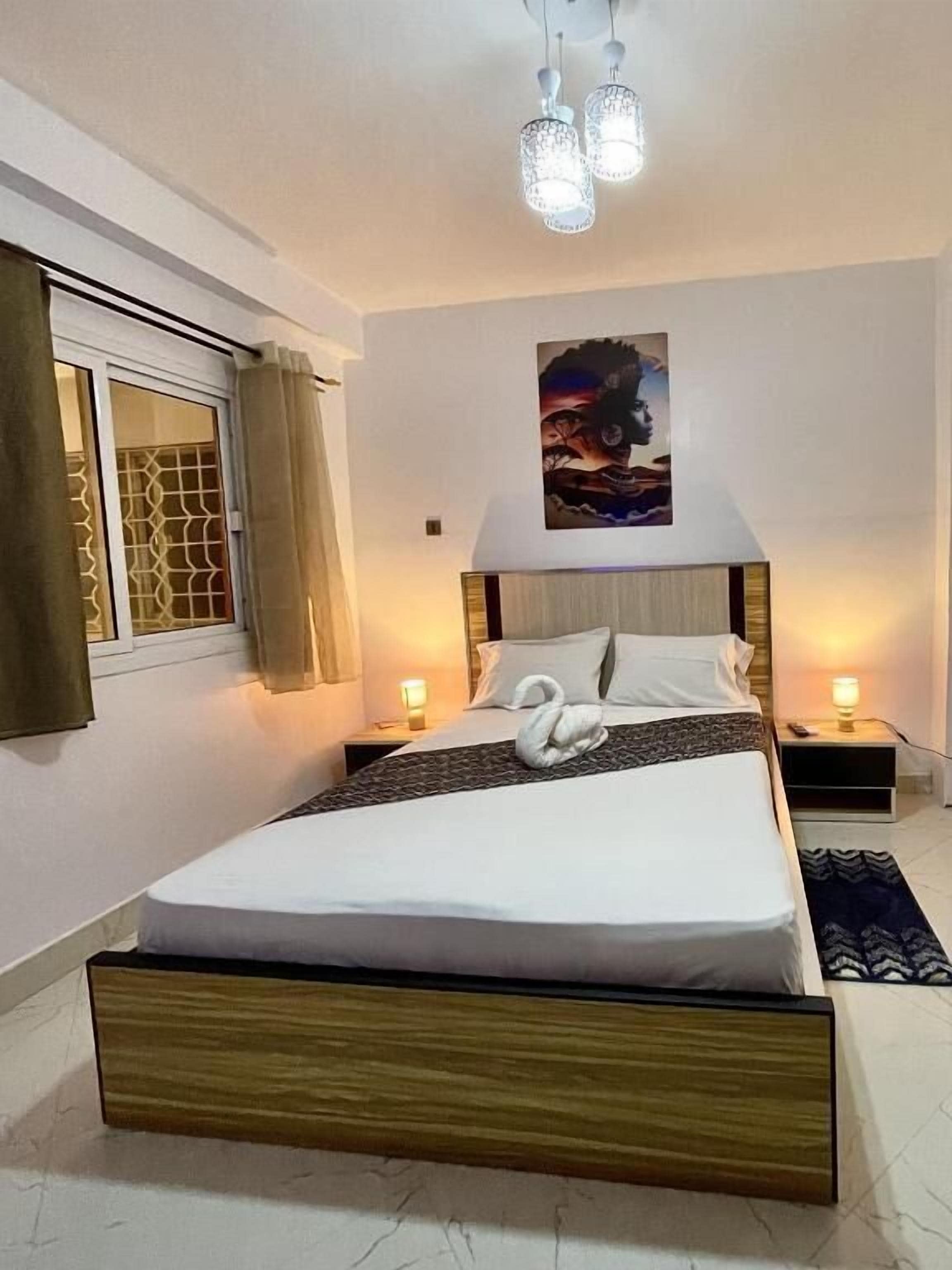 A bedroom with a wooden bed, two bedside tables, a window with curtains, a painting on the wall, and a chandelier.
