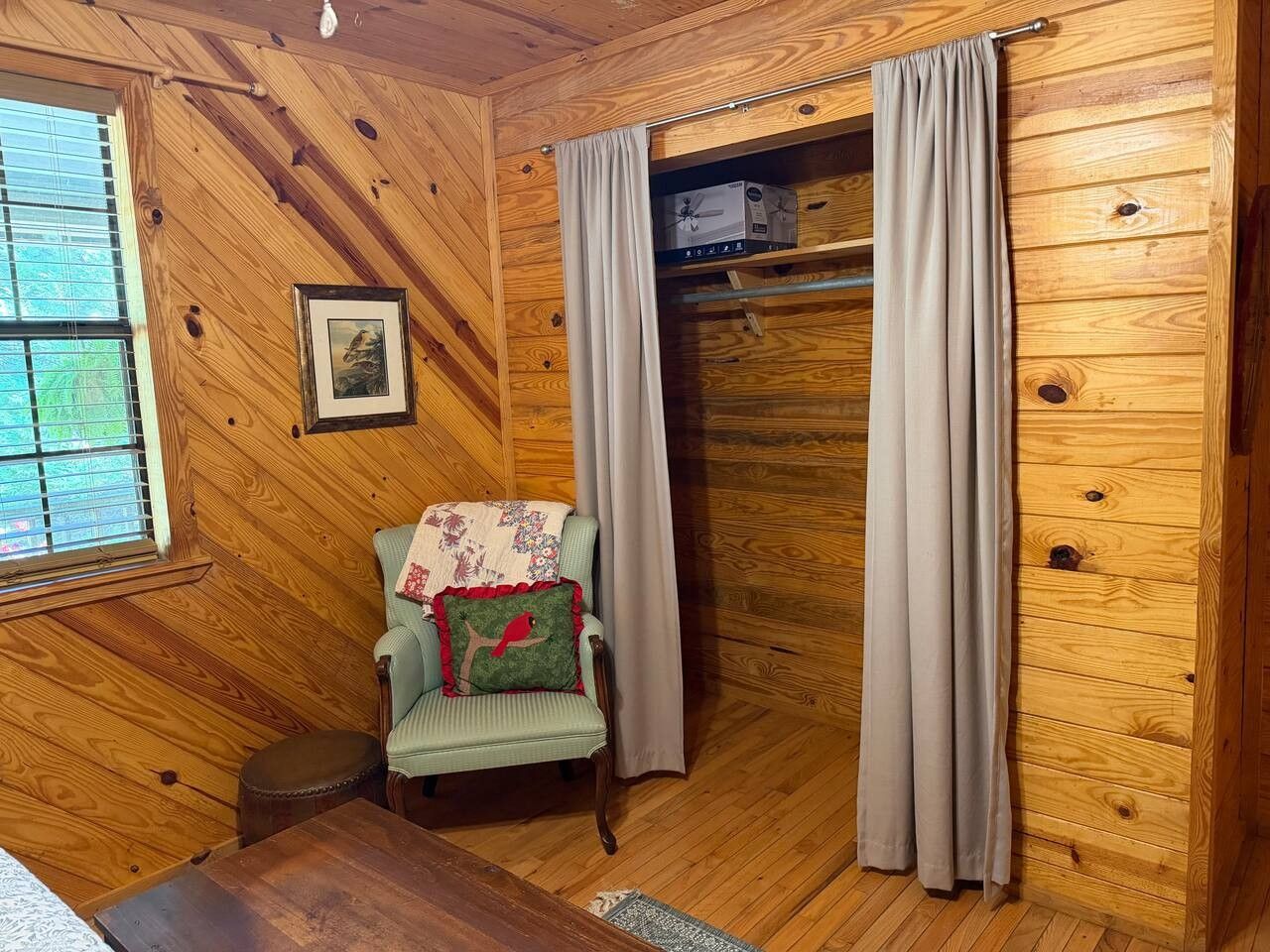 A wooden-paneled room with a chair, a small table, a window with blinds, and a door with curtains.