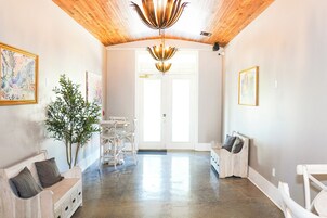Un pasillo espacioso con techo de madera, una lámpara de araña grande y una puerta que da a otra habitación. Hay dos sofás blancos con cojines, una planta en maceta y obras de arte en las paredes.