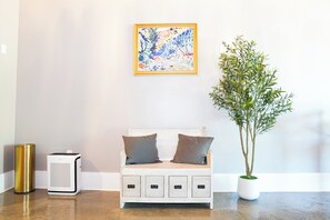 A room with a white sofa, two pillows, a framed painting, a potted plant, and a white speaker.