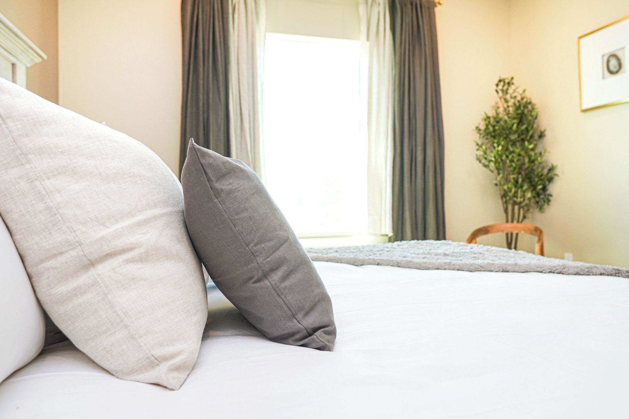 Un lit avec deux oreillers, une fenêtre avec des rideaux, une chaise et une plante dans une chambre d’hôtel.