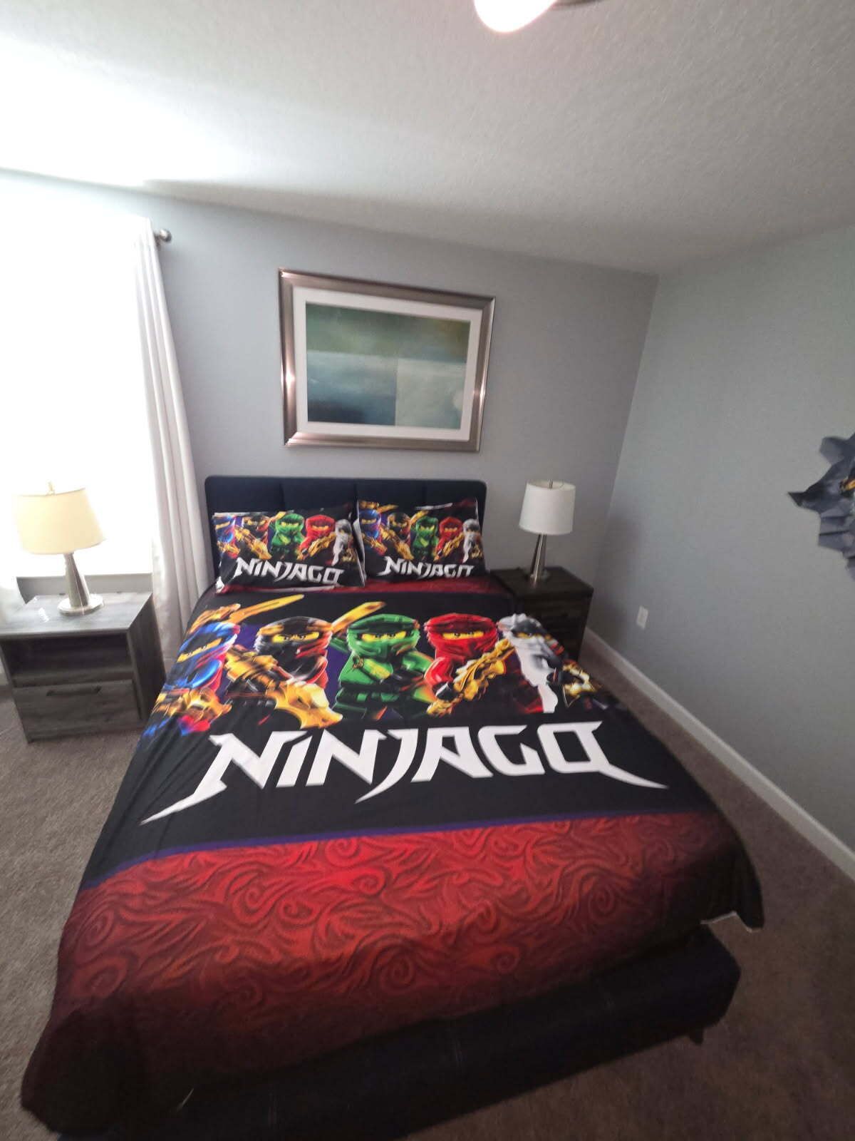 Un dormitorio con una cama y un edredón con temática de Ninjago, un cuadro enmarcado en la pared y una mesita de noche con una lámpara.