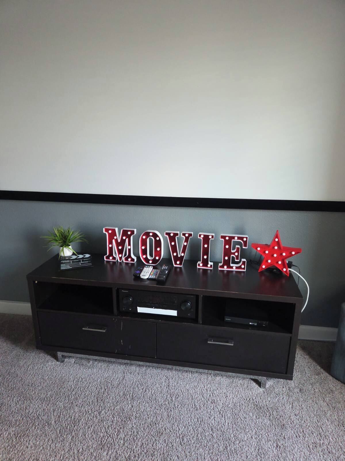 Eine dunkle Holz-Unterhaltungsecke mit der leuchtenden Aufschrift 'MOVIE', einer kleinen Topfpflanze und einer roten Stern-Deko.