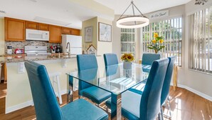 A modern kitchen with wooden cabinets, a microwave, and a refrigerator. There is a dining area with a glass table and blue chairs. - 3BR family condo | steps from Laguna Bay pool (Kissimmee)