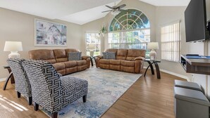 A living room with two brown sofas, patterned ottoman, and a ceiling fan. - 3BR family condo | steps from Laguna Bay pool (Kissimmee)