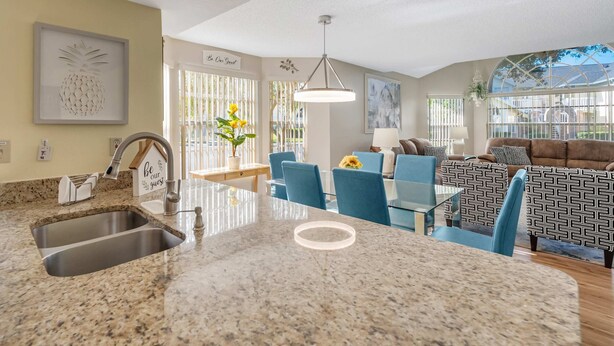 A modern kitchen with granite countertops, a built-in sink, and a dining area with blue chairs and a patterned sofa. - 3BR family condo | steps from Laguna Bay pool (Kissimmee)