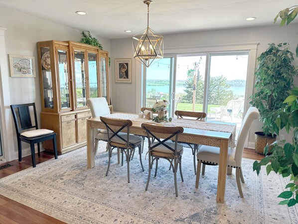 A dining area with a wooden table, chairs, a chandelier, and large windows with a view of the outdoors.
