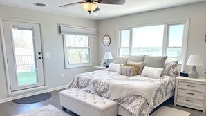 Un dormitorio con cama, una banca, mesita de noche, lámpara, ventilador de techo y vistas al exterior.