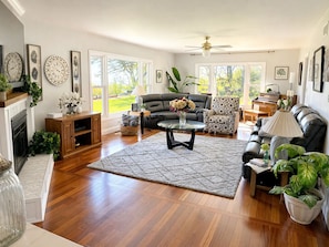 A spacious living room with hardwood flooring, a fireplace, a large window, a ceiling fan, and a variety of furniture including a sofa, armchairs, and a coffee table.
