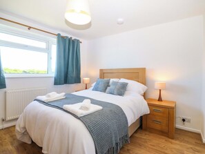 A neatly made bed with a headboard, bedside lamps, a window with curtains, and a radiator. - 20 Gwelfor Estate (Cemaes Bay)
