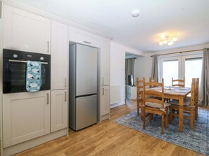 A compact kitchen with a refrigerator, oven, and a dining table with chairs. - 20 Gwelfor Estate (Cemaes Bay)