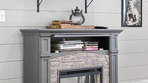 Een open haard met een stenen gevel, een houten schouw met boeken en een vintage lamp, en een ingelijst schilderij aan de muur.