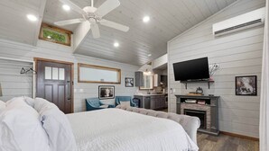 Een slaapkamer met een groot bed, een plafondventilator, een televisie, een open haard en een keuken in de achtergrond.