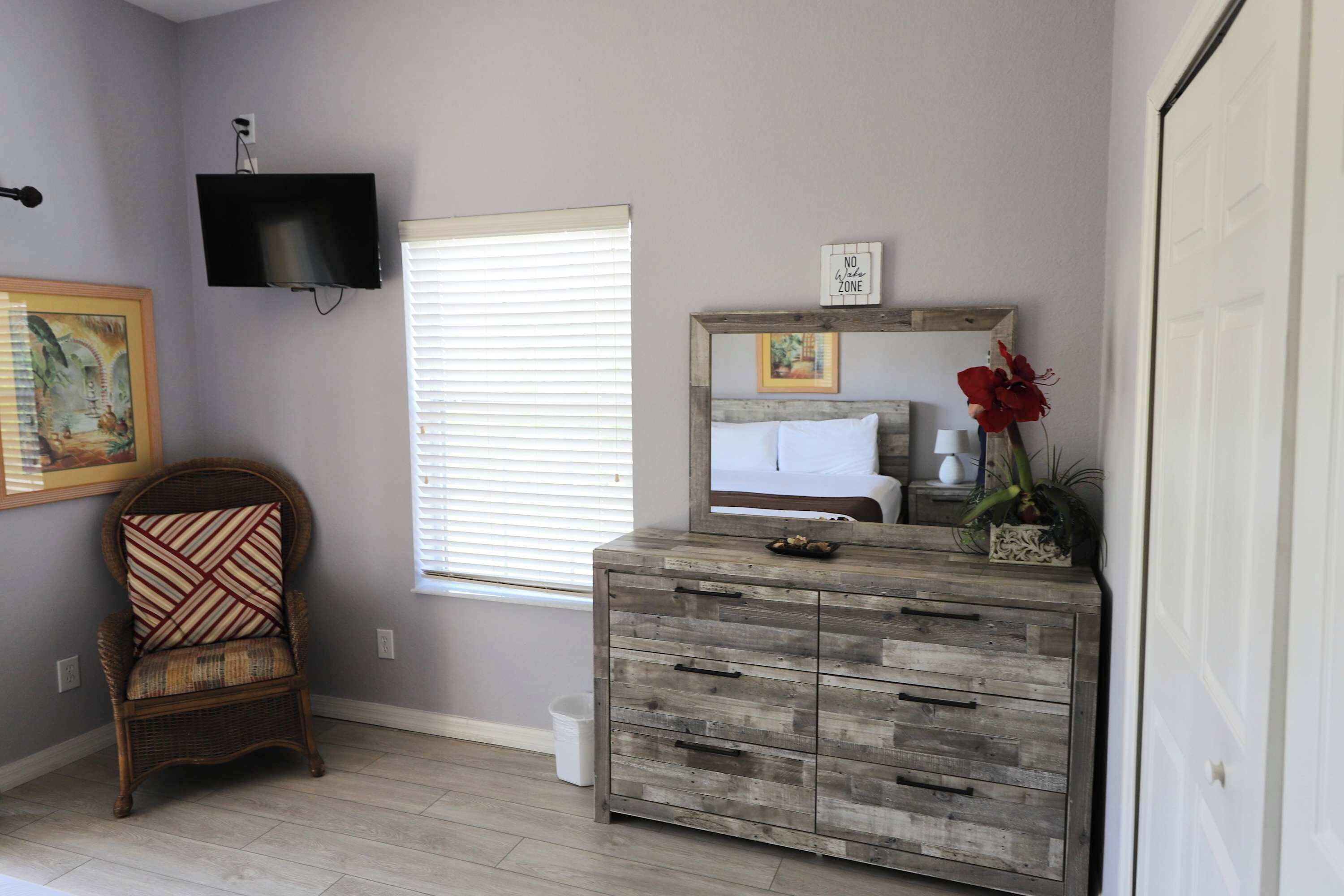A room with a wooden dresser, a chair, a television mounted on the wall, and a window with blinds.