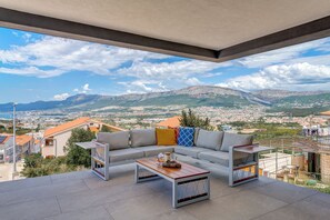 A modern outdoor seating area with a sofa and coffee table, overlooking a cityscape and mountains. - Panorama Coastal Haven (Split)