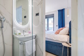 Un baño con lavamanos de mármol, cabina de ducha de vidrio y un espejo. La habitación tiene una cama con cortinas azules y una mesita de noche con un control remoto.