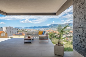 En modern utomhusplats med en soffa, ett soffbord och en krukvÀxt, med utsikt över en stadssiluett och berg i fjÀrran.