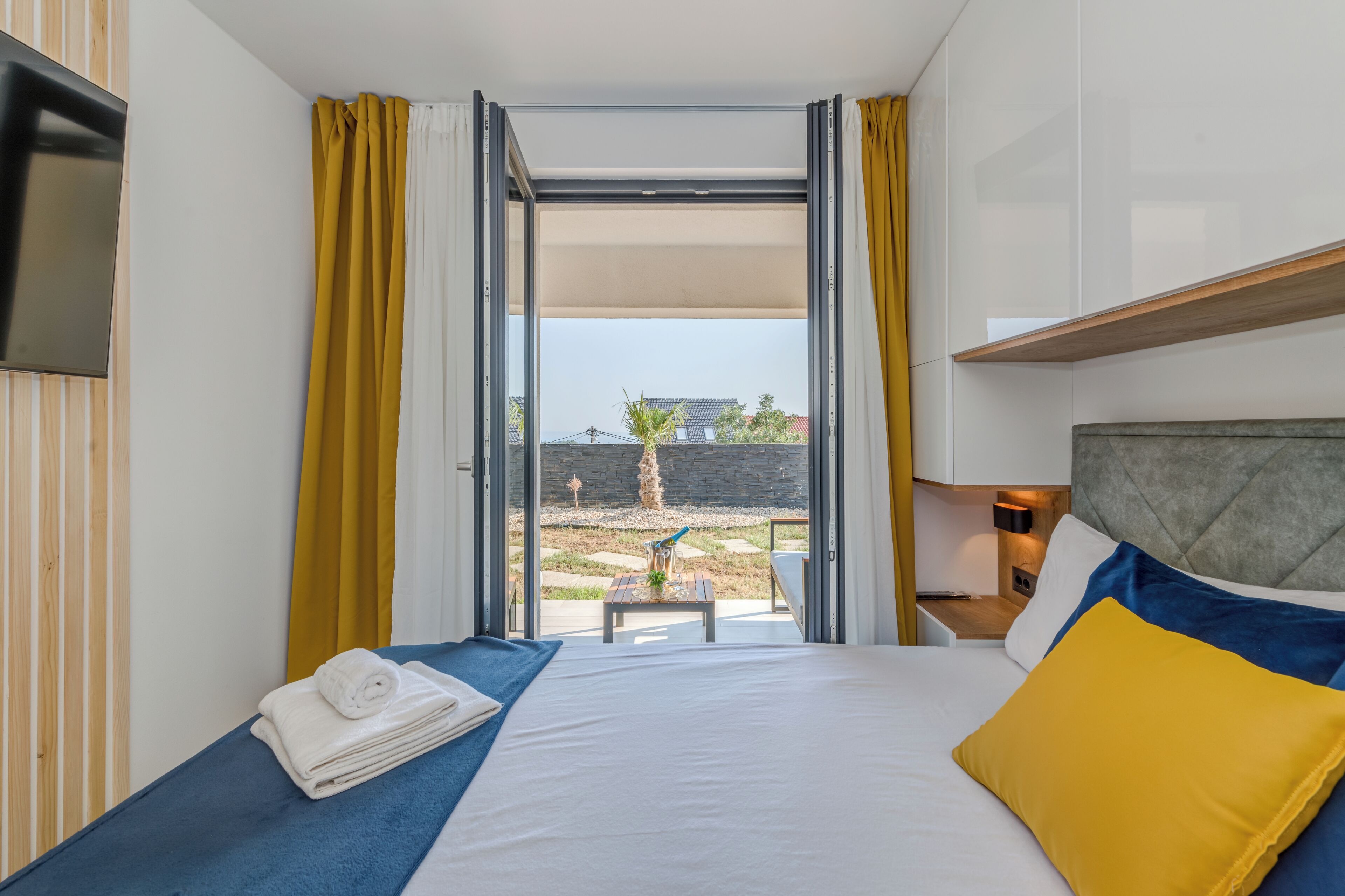 A hotel room with a bed, a TV mounted on the wall, a small table with a plant, and a view of the outside.