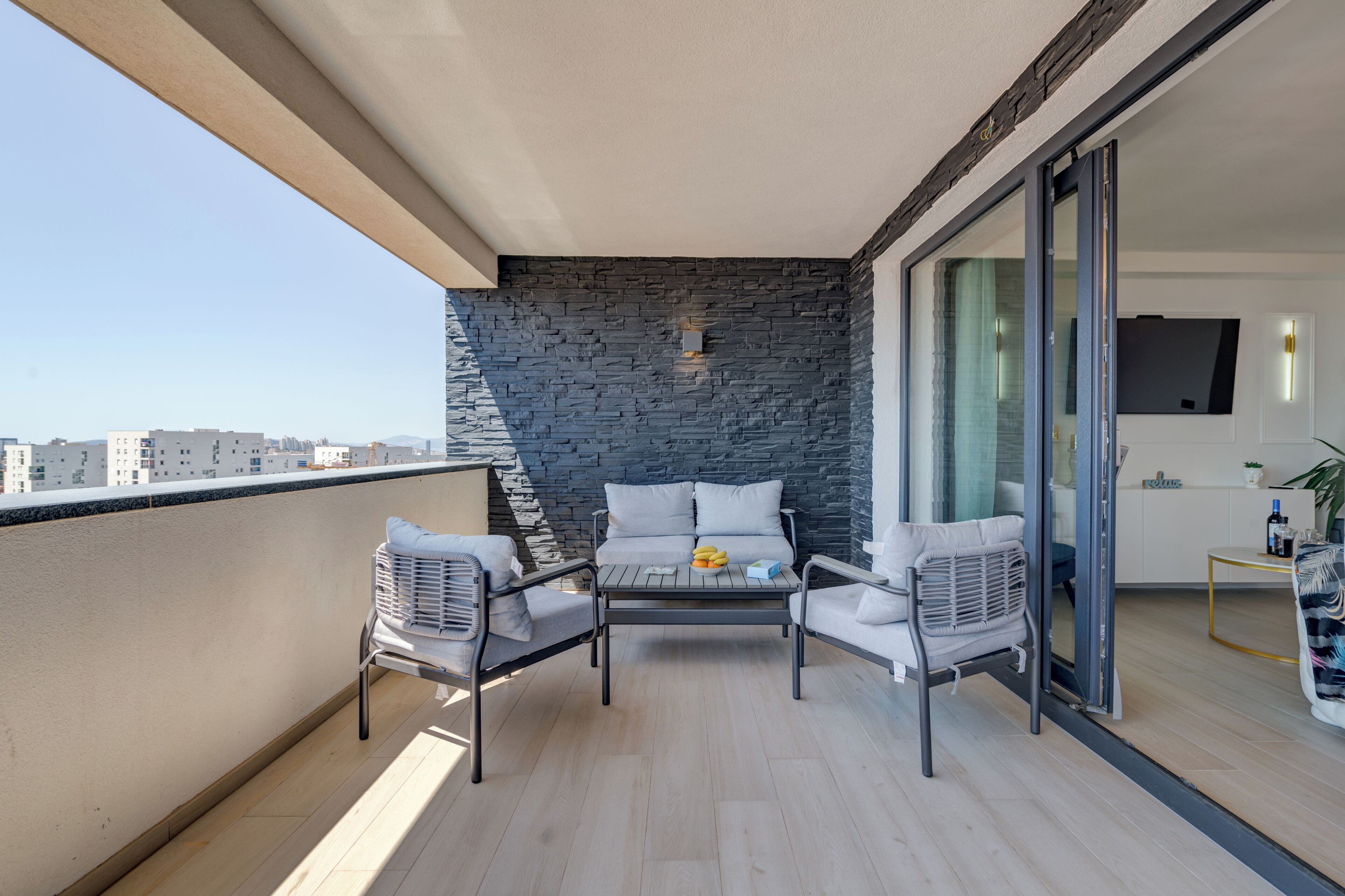 A balcony with a stone wall, a sofa, two chairs, a table with fruit, and a sliding glass door leading to an interior space.