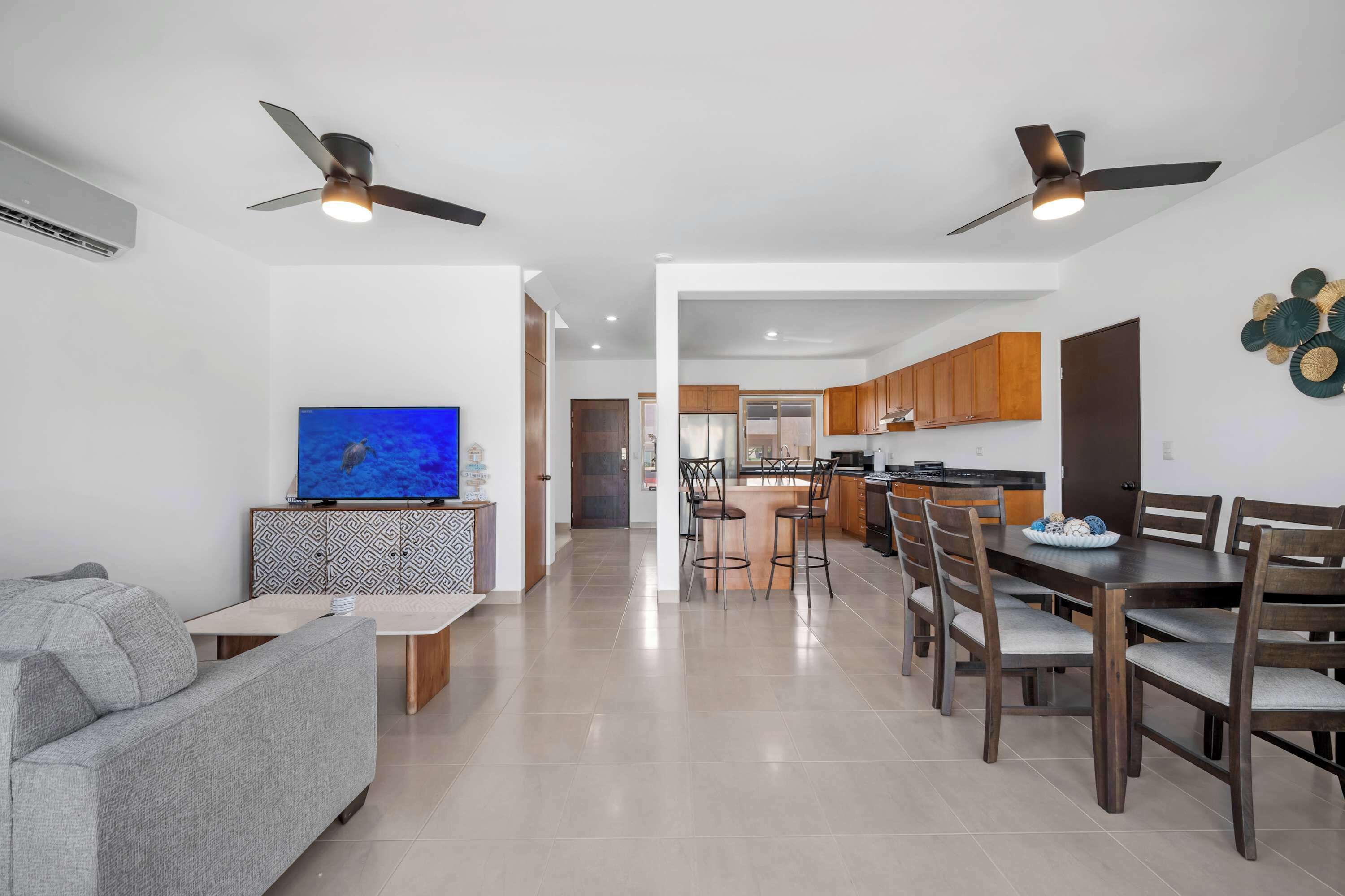 Una sala de estar moderna con televisor, sofá, mesa de comedor, sillas, ventiladores de techo y una cocina al fondo.