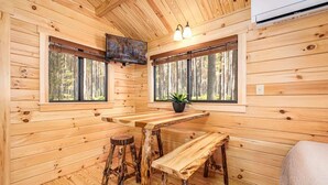 A wooden cabin interior with a wooden table, stools, a potted plant, and two windows with a forest view.