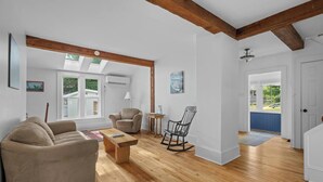 Ein geräumiges Wohnzimmer mit Holzboden, einer Couch, einem Schaukelstuhl und einem Tisch. Ein Dachfenster und eine Tür zum Garten sind vorhanden.