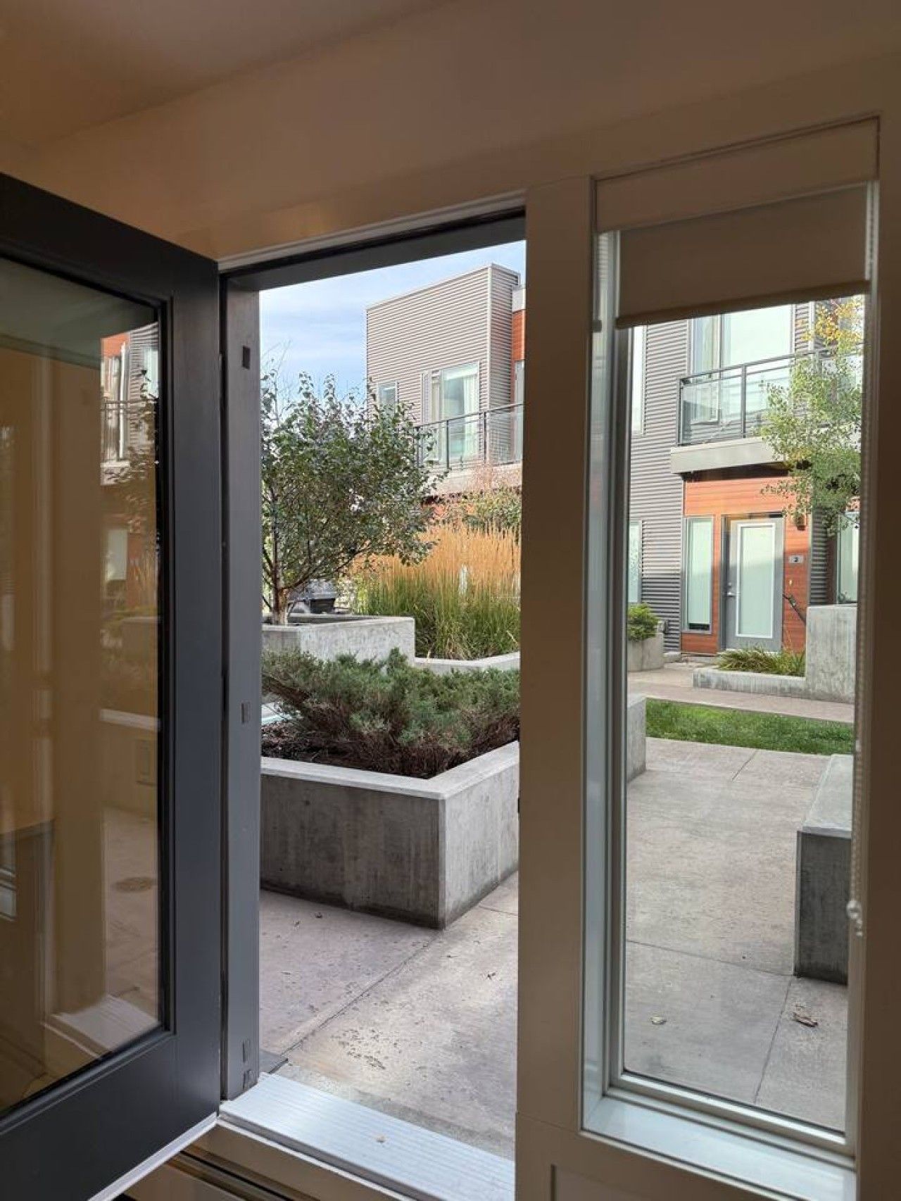 A view through a window of a courtyard with a modern building, a concrete planter with shrubs, and a clear sky.