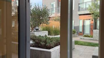 A view through a window of a courtyard with a modern building, a concrete planter with shrubs, and a clear sky.
