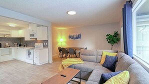 A modern living room with a sofa, a dining table with chairs, a kitchen area with white cabinets and appliances, and a large window with curtains. - Cozy 2 Bedroom Suite Near Ctrain Mall and Downtown (Calgary)
