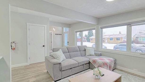 A living room with a grey sofa, a coffee table with a vase, and large windows with a view of a parking lot and buildings.