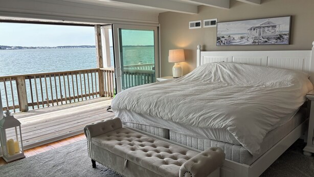 A bedroom with a large bed, a bench, a nightstand with a lamp, and a view of the water.