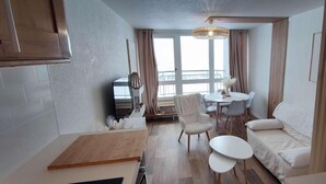 Ett kompakt bostadsutrymme med ett litet kök, en matplats med ett runt bord och stolar, en soffa och ett stort fönster som släpper in naturligt ljus.