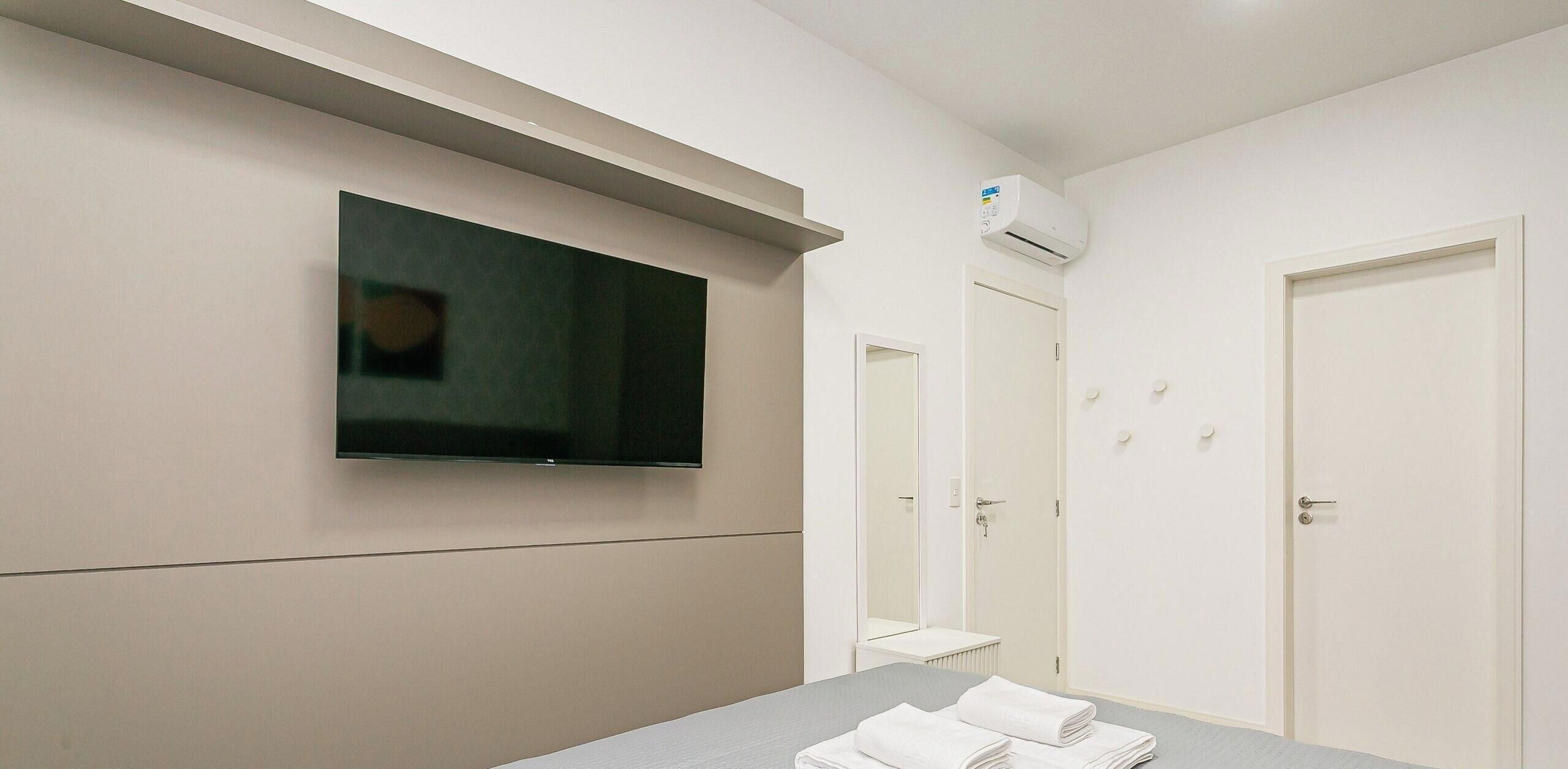 Una camera d'albergo con televisore a schermo piatto montato a parete, un letto con asciugamani bianchi piegati con cura, una porta bianca e una piccola finestra.