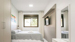 A bedroom with a bed, a TV mounted on the wall, a window with curtains, and a wardrobe. - Apartamento Águas da Cachoeira - FLN (Florianópolis)
