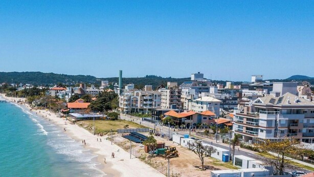 A coastal cityscape with a beach, buildings, and a distant mountain. - Apartamento Águas da Cachoeira - FLN (Florianópolis)