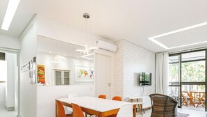 A modern living room with a dining area, a flat-screen TV mounted on the wall, and a large window with curtains. - Apartamento Águas da Cachoeira - FLN (Florianópolis)