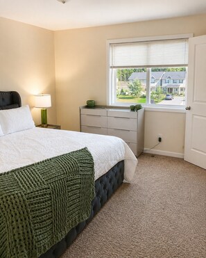 Un dormitorio con cama, lámpara de noche, cómoda, ventana con persianas y vistas a casas.