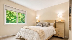A bedroom with a bed, two bedside tables with lamps, a window with a view of trees, and a carpeted floor. - Holiday Village Townhomes – 3883 (Traverse City)