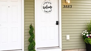 A house with two white doors, a welcome sign, a house number 3883, a small potted plant, and a doormat with 'hello' on it. - Holiday Village Townhomes – 3883 (Traverse City)