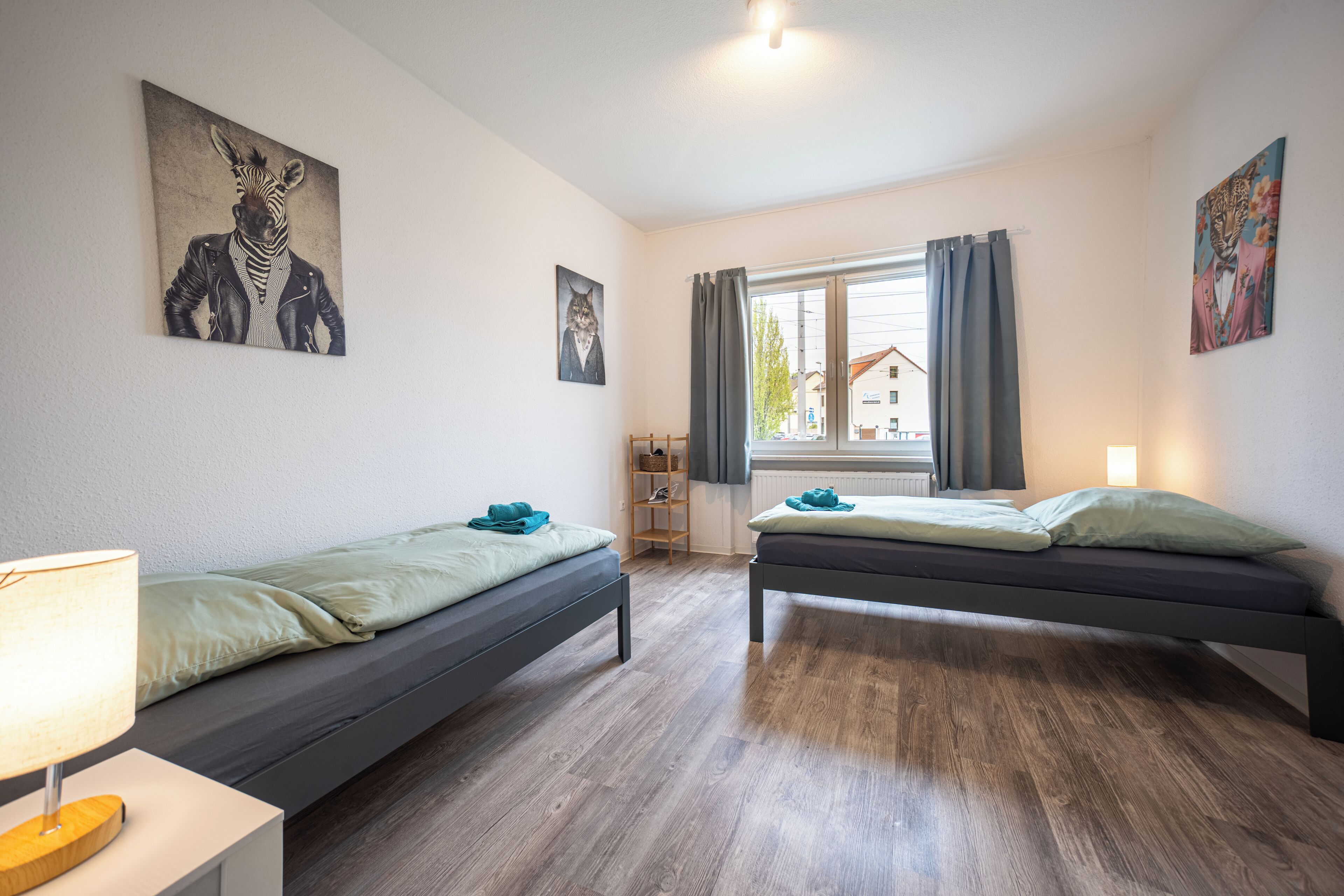 Et værelse med to senge, et natbord med lampe, et vindue med gardiner og billedkunst på væggene.